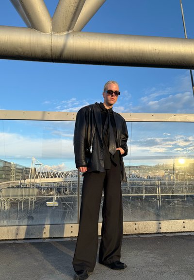 Blazer oversize de cuero negro, camisa negra y pantalones de pierna ancha. La modelo lleva gafas de sol frente a un fondo de cielo azul y estructuras metálicas.