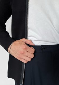 Juodas megztinis su žakardo raštu ir pilkos spalvos užtrauktuku, dėvimas ant balto marškinėlio, o ranka laiko tamsiai mėlynas kelnes su sagčių uždarymu.