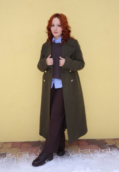 Mujer con cabello rizado rojo, vestida con un abrigo largo verde, camisa azul y pantalones oscuros, de pie contra una pared amarilla sobre un suelo de baldosas nevado.