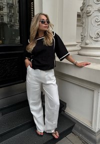 Sort linned top med creme trim, brede hvide linnedbukser og sorte flip-flops. Står på sorte trin nær en udsmykket hvid søjle.
