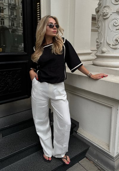 Top de lino negro con ribete crema, pantalones anchos de lino blanco y chanclas negras. De pie en unos escalones negros cerca de una columna blanca ornamental.