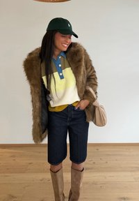 Γυναίκα που χαμογελά με μακριά σκούρα μαλλιά, φορώντας πράσινο καπέλο, πολύχρωμο πουκάμισο, παλτό από γούνα, τζιν σορτς, ταμπά μπότες και κρατώντας μια μπεζ τσάντα.