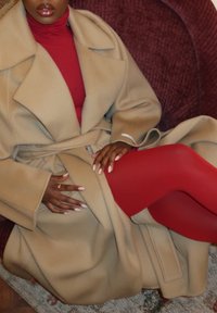 Abrigo de lana beige con solapas anchas y cinturón, combinado con un jersey de cuello alto rojo y medias a juego. Texturas suaves y diseño ajustado.