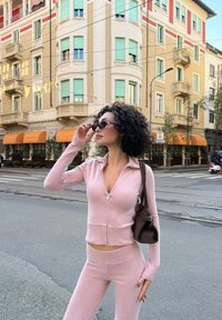 Jasnoróżowy sweter na zamek i dopasowane spodnie z miękkiego materiału, zestawione z ciemnymi okularami przeciwsłonecznymi i brązową torbą na ramię. Miejski krajobraz w tle.