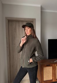 Γυναίκα με πράσινο-ελιά μπουφάν και καπέλο που ταιριάζει, που κλείνει το μπουφάν της με φερμουάρ, στέκεται μέσα σε κλειστό χώρο κοντά σε ξύλινη πόρτα και ντουλάπι με τηλεόραση από πάνω.