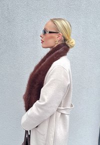 Kreemjas villane mantel rihmaga, pruun kasuka sall, mustad päikeseprillid, kuldsed spiraalkõrvarõngad, siledad juuksed ülespandud kuklasse, tekstuuriga hall taust.