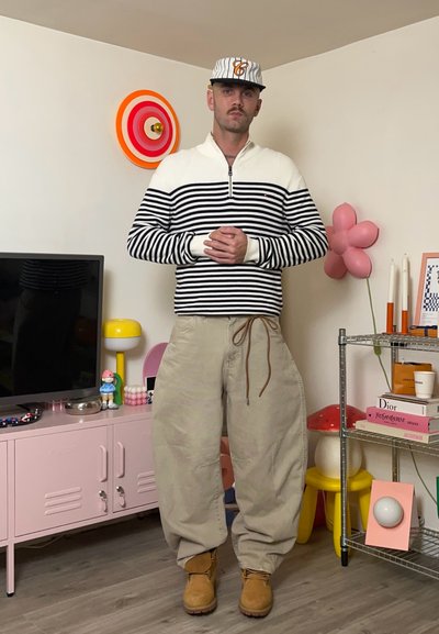 Suéter de rayas de manga larga en blanco y azul marino, pantalones beige holgados y botas marrones claras. Los accesorios incluyen una gorra y un reloj. Decoración colorida en el fondo.