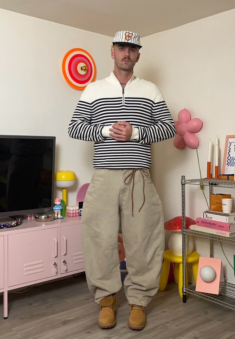 Maglione a righe con maniche lunghe bianco e blu navy, pantaloni beige larghi e stivali color cuoio. Accessori includono un cappellino e un orologio. Decorazioni colorate sullo sfondo.