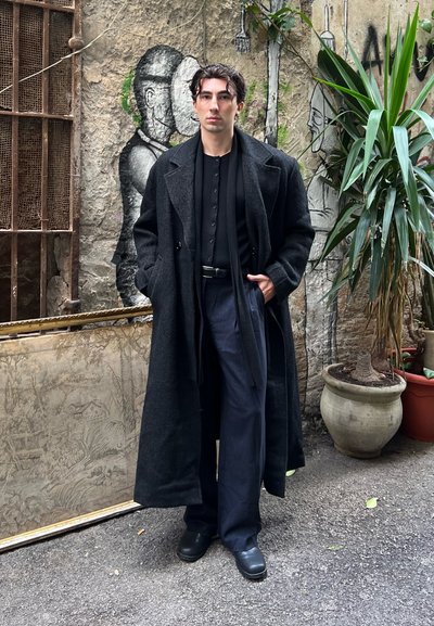 Abrigo largo de lana negra sobre una camisa negra abotonada, combinado con pantalones oscuros de rayas finas y zapatos negros. El fondo presenta vegetación y un mural.