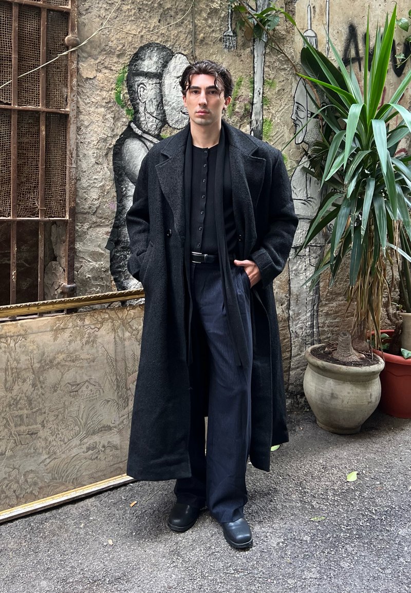 Long manteau noir en laine porté sur une chemise noire boutonnée, associé à un pantalon sombre à fines rayures et des chaussures noires. En arrière-plan, on aperçoit de la verdure et une fresque murale.