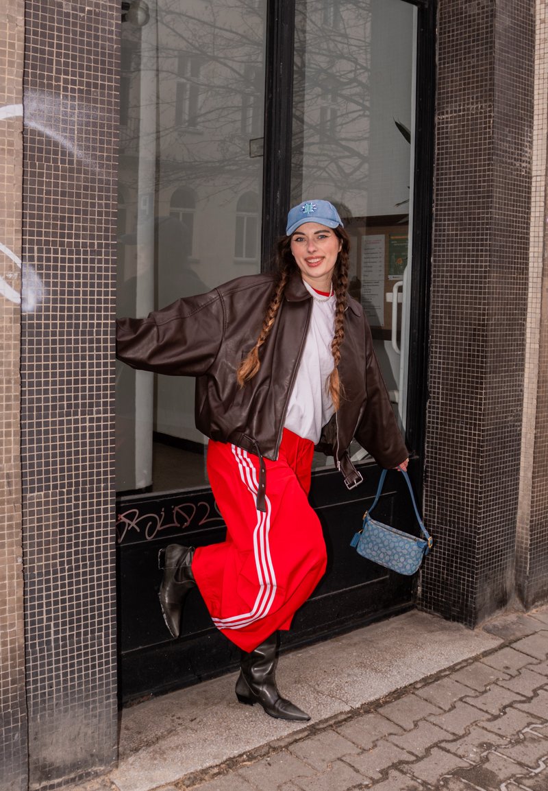 Jeune femme aux cheveux tressés portant une casquette bleue, une veste en cuir marron, une chemise blanche, un pantalon rouge, des bottes noires, et tenant un sac à main bleu, debout près d'une porte.