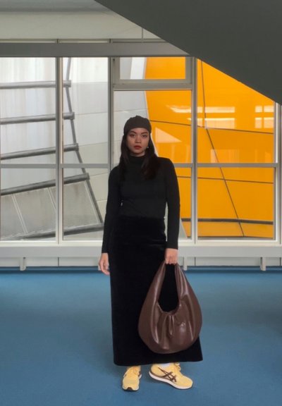 Suéter de cuello alto negro, falda maxi de terciopelo negro, bolso hobo marrón y zapatillas amarillas. El fondo cuenta con grandes ventanales y una pared naranja.