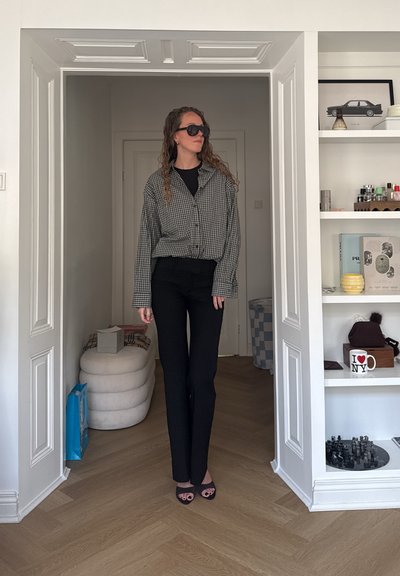 Mujer con cabello largo y rizado, con gafas de sol, chaqueta a cuadros, pantalones negros y sandalias, de pie en un marco de puerta blanco cerca de estanterías con decoración.