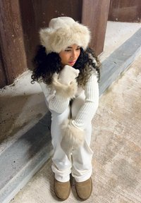 Maglione bianco a coste e pantaloni, guanti e cappello con accenti di pelliccia, stivali in vernice marrone, capelli ricci scuri, seduto contro un muro testurizzato.