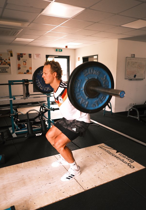 Man hurkt met een geladen halter op zijn schouders binnen een sportschool met gewichthefapparatuur.