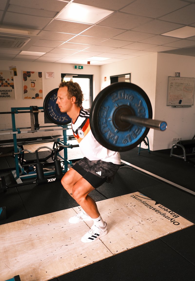 Mand squatter med en belastet vægtstang på skuldrene inde i et fitnesscenter med vægtløftningsudstyr.