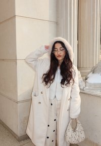 Γυναίκα με μακριά σκούρα μαλλιά, ντυμένη με λευκό παλτό με κουκούλα και πουά παντελόνι, κρατώντας μια λευκή τσάντα διακοσμημένη με μαργαριτάρια δίπλα σε ένα κτίριο με κίονες.