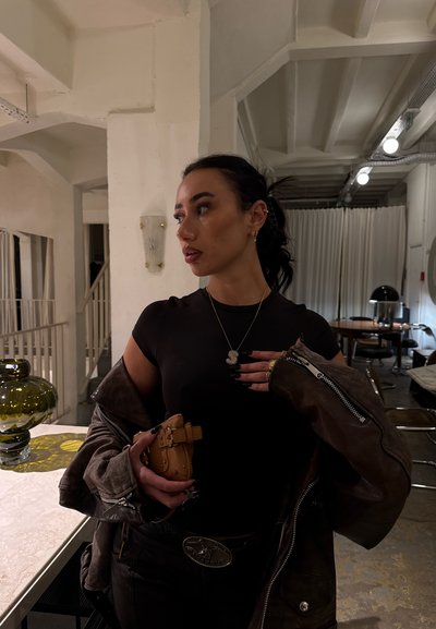 Mujer de pelo oscuro recogido, vestida con una blusa negra y una chaqueta marrón, sosteniendo un pequeño bolso beige, de pie en una habitación moderna con mesas y sillas.