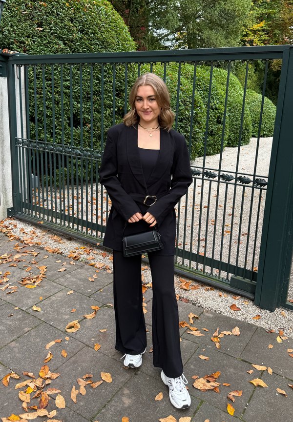 Blazer preto alfaiatado com cinto, combinado com uma blusa preta e calças pretas de perna larga. Ténis brancos desportivos e uma pequena mala preta.