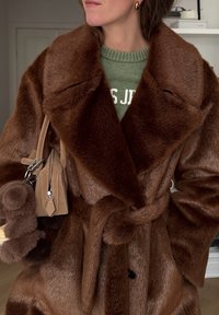 Femme portant un manteau en fausse fourrure marron noué à la taille sur un pull vert, portant un sac à main en daim beige avec des porte-clés en peluche.