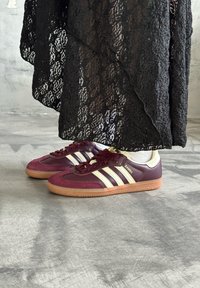 Bordeauxmockaskor med tre vita ränder och gummisula; bärs med ett strukturerat svart spetsplagg; visar spetsmönsterdetaljer.