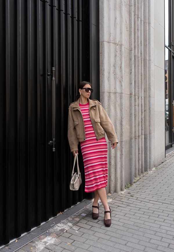 Frau mit Sonnenbrille, die eine braune Jacke und ein rot gestreiftes Kleid trägt, lehnt an einer schwarzen Metallwand auf einem Bürgersteig in der Stadt.