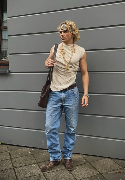 Camisa sin mangas color bronce, jeans de mezclilla azules, bolso de hombro marrón, collar decorativo, cabello rizado rubio, botas marrones con estampado, de pie contra una pared gris.