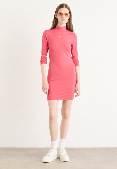 Vestido ajustado rosa con cuello alto y mangas largas, hecho de un material suave. Usado con zapatillas blancas y calcetas hasta el tobillo.