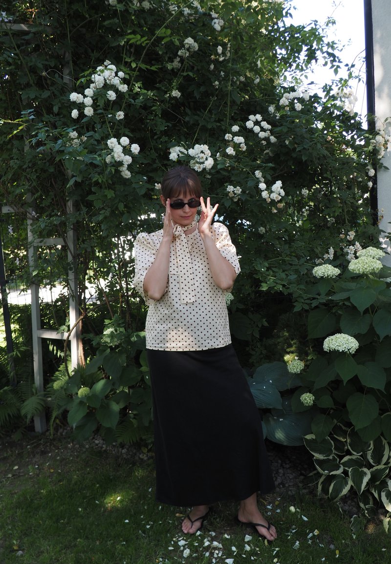 Femme portant des lunettes de soleil, un chemisier à pois et une jupe noire, debout pieds nus dans un jardin entouré de plantes à fleurs blanches et de feuillage vert.