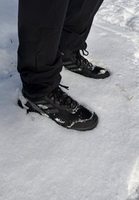 Bottes d'hiver noires avec de la neige dessus, portées avec un pantalon noir, debout sur de la neige fraîche à l'extérieur.