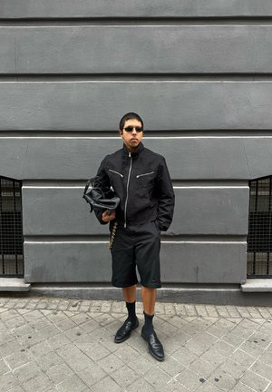 Man wearing a black jacket, shorts, sunglasses, holding a black bag, standing on the pavement against a grey wall with two small barred windows.