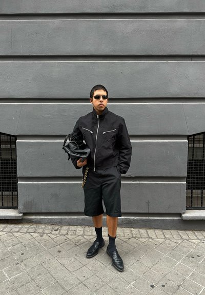 Hombre con chaqueta negra, pantalones cortos, gafas de sol, sosteniendo una bolsa negra, de pie en la acera frente a una pared gris con dos pequeñas ventanas con rejas.