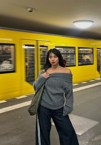 Grijze geribde trui met blote schouders gecombineerd met losse donkere spijkerbroek; een groene boodschappentas in de hand. Gele metrotrein zichtbaar op de achtergrond.