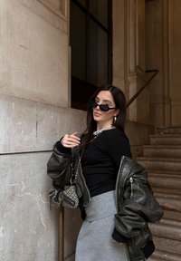 Υπερμεγέθη ελαιοπράσινη μπόμπερ ζακέτα, εφαρμοστό μαύρο τοπ, γκρι πλεκτή φούστα, μαύρα γυαλιά ηλίου, κολιέ με χάντρες και τσάντα με σχέδιο. Φόντο με πέτρινο τοίχο.