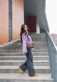 Col roulé tricoté violet, jean évasé gris, bottes à plateforme couleur cuir clair et sac à main marron texturé. Debout sur des escaliers extérieurs avec des murs en arrière-plan.