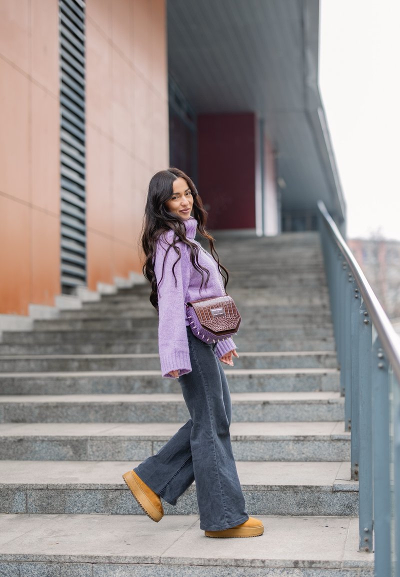Lila gestrickter Rollkragenpullover, graue ausgestellte Jeans, braune Plateaustiefel und eine strukturierte braune Handtasche. Steht auf einer Außentreppe mit Wänden im Hintergrund.