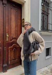 Χοντρή μπεζ μπλούζα, γκρι κασκόλ, και μαύρη δερμάτινη τσάντα; χαλαρώνοντας δίπλα σε μια ξύλινη πόρτα με λεπτομερείς χαρακιές; γκρι τοίχος και παράθυρο με κάγκελα.