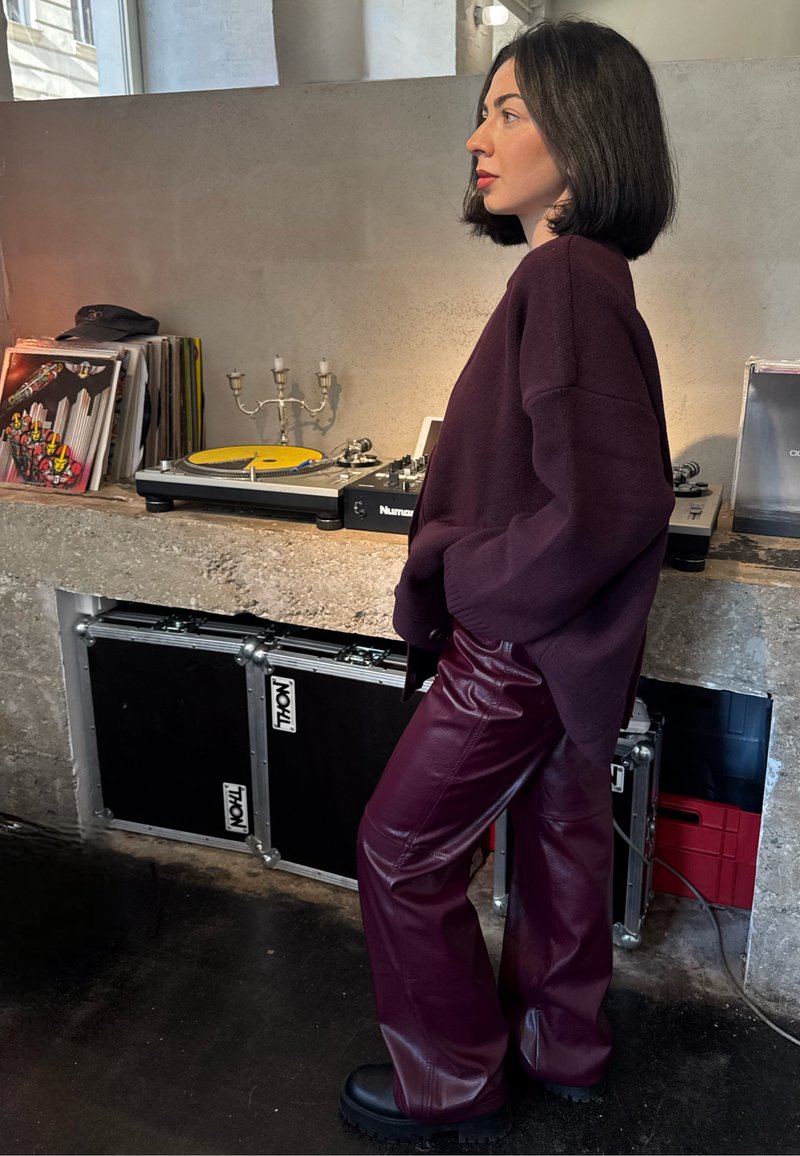 Sweater oversized em malha cor bordô, combinada com calças de cintura alta em pele sintética de perna larga. Botas pretas robustas. Fundo com discos de vinil e equipamento.