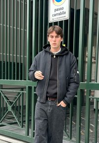 Veste zippée sombre avec capuche, portée par-dessus un pull noir, associée à un jean noir ample. Debout devant une porte verte.