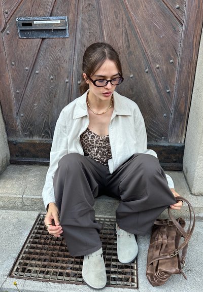 Camisa de lino crema sobre una blusa de estampado de leopardo, pantalones de pierna ancha marrones, zapatos de ante grises, sosteniendo un bolso de cuero marrón, sentado cerca de una puerta de madera.