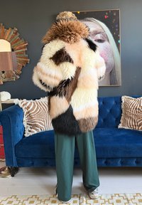 Personne portant un manteau en fourrure duveteuse multicolore et un pantalon vert foncé, faisant face à un canapé bleu avec des coussins à motifs dans un salon moderne.