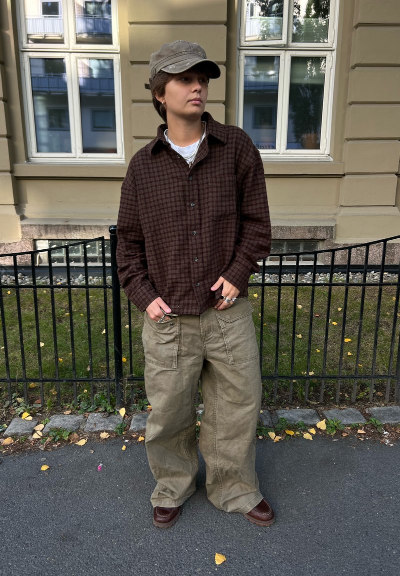 Person med brun rutete skjorte, beige vide bukser, brune sko og en grå caps som står på fortauet nær et svart metallgjerde og en beige bygning.