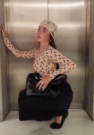 Blusa de manga larga con patrón de luna creciente, color beige; bolso negro de estilo relajado, y zapatos de tacón negros. Gorro de crochet y anillos visibles.