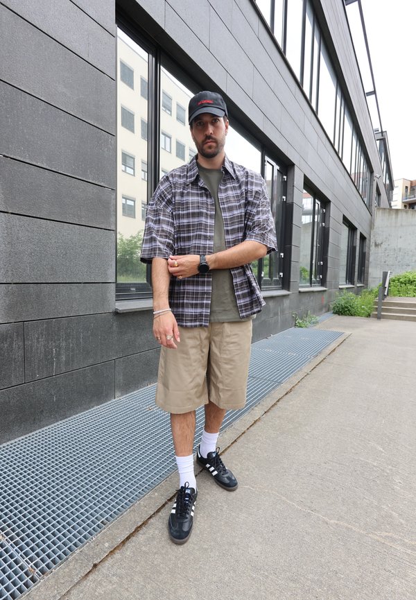 Mann mit kariertem Hemd, khakifarbenen Shorts, schwarzer Kappe und schwarzen Sneakers steht auf einem städtischen Gehsteig nahe einem modernen Gebäude mit großen Fenstern.