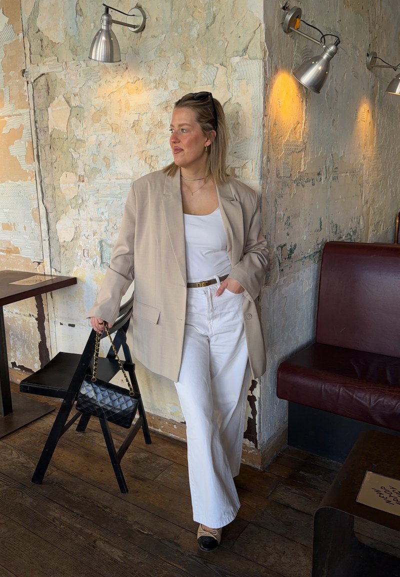 Femme en blazer beige, top blanc et pantalon, appuyée contre un mur texturé dans un café rustique, tenant un sac à main noir avec une chaîne dorée.