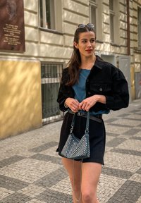 Melna pārāk liela jaka, tirkīza topiņš, augsto jostasvietu melnas šortus un maza trīsstūrveida pērlīšu soma ar ķēdes siksnu. Redzams ietves raksts.