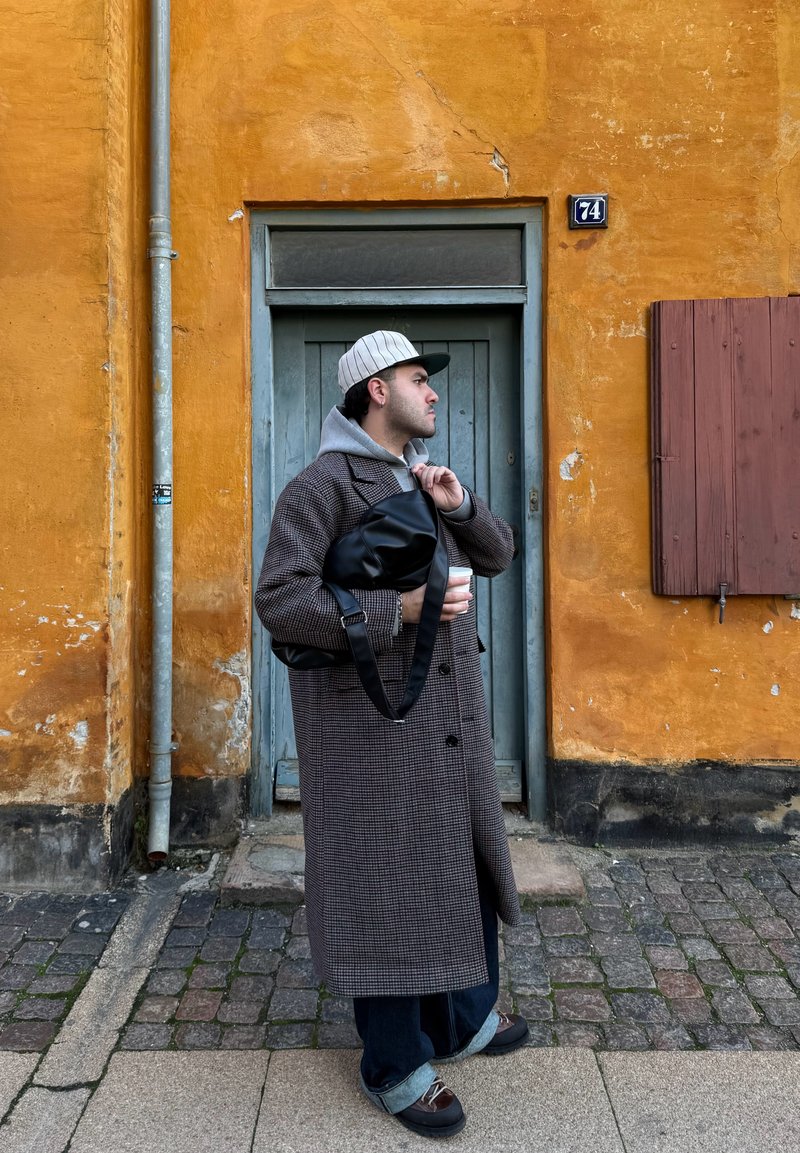 Homme portant un long manteau à carreaux, un sweat à capuche gris et une casquette rayée, tenant un sac noir et une tasse de café, debout près d'un mur orange usé avec une porte bleue.