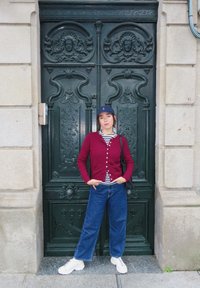 Cardigã bordô com fecho de botão sobre uma camisa riscada, combinado com jeans de perna larga azuis e ténis brancos, contra uma porta verde ornamentada.