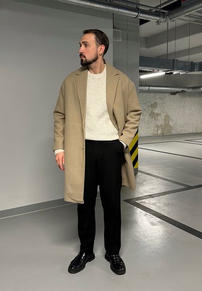 Abrigo beige sobre un suéter blanco texturizado, combinado con pantalones negros y zapatos negros brillantes, en un entorno minimalista de tonos grises y concreto.