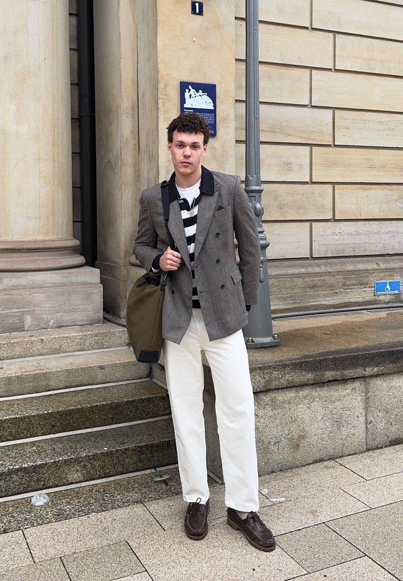 Young man with curly hair stands by stone steps and pillar, wearing a grey blazer, striped shirt, white trousers, and holding an olive bag.
